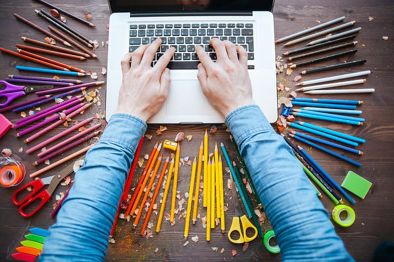 Een persoon die op een laptop werkt met kleurpotloden in de buurt.